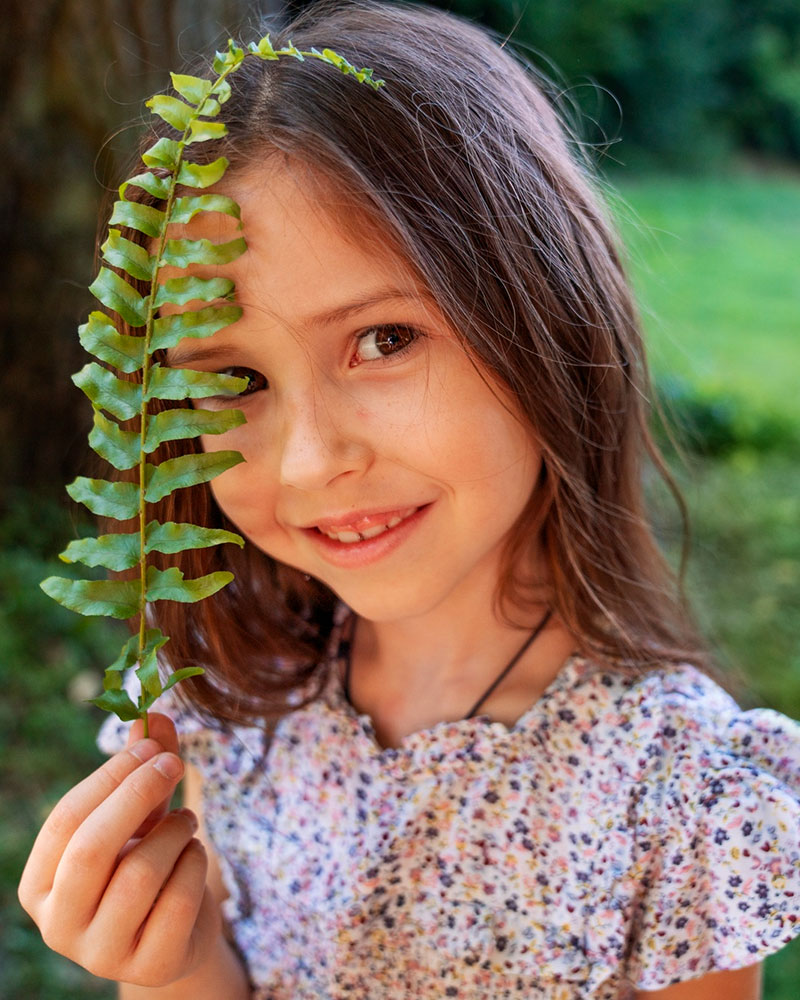 A girl holds up a fern leaf next to her face, she is looking at the camera and smiling.
